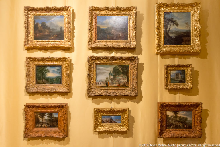 Nine small landscape paintings in ornate gilded frames arranged closely on a pale wall.