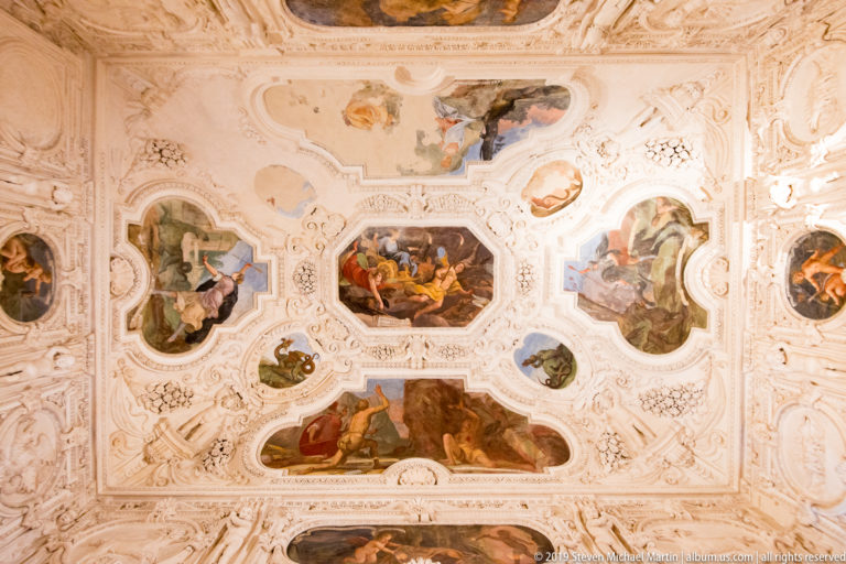 Ornate white stucco ceiling framing multiple painted figurative scenes in irregular panels.