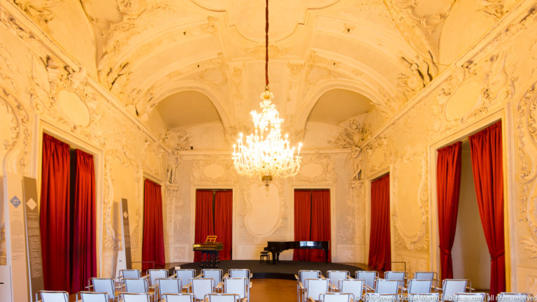 White stucco salon with red curtains, crystal chandelier, rows of chairs, and a keyboard instrument.