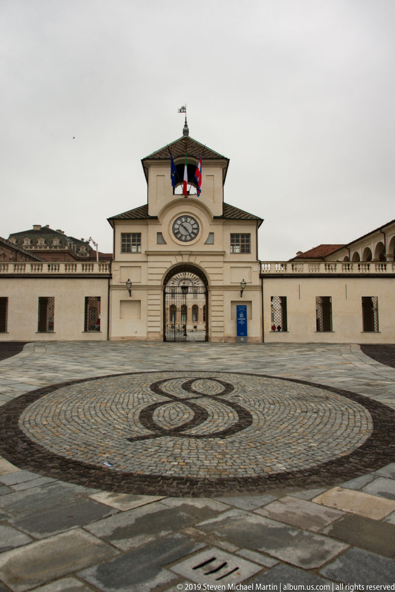 Clock gate with flags beyond a large circular black-and-white cobblestone motif in the forecourt of La Venaria Reale.