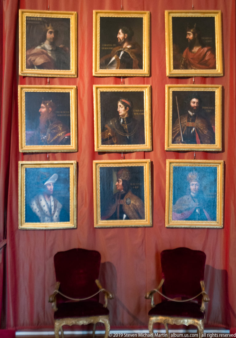 Nine gilded framed historical portraits arranged in a grid on a red wall above two chairs.