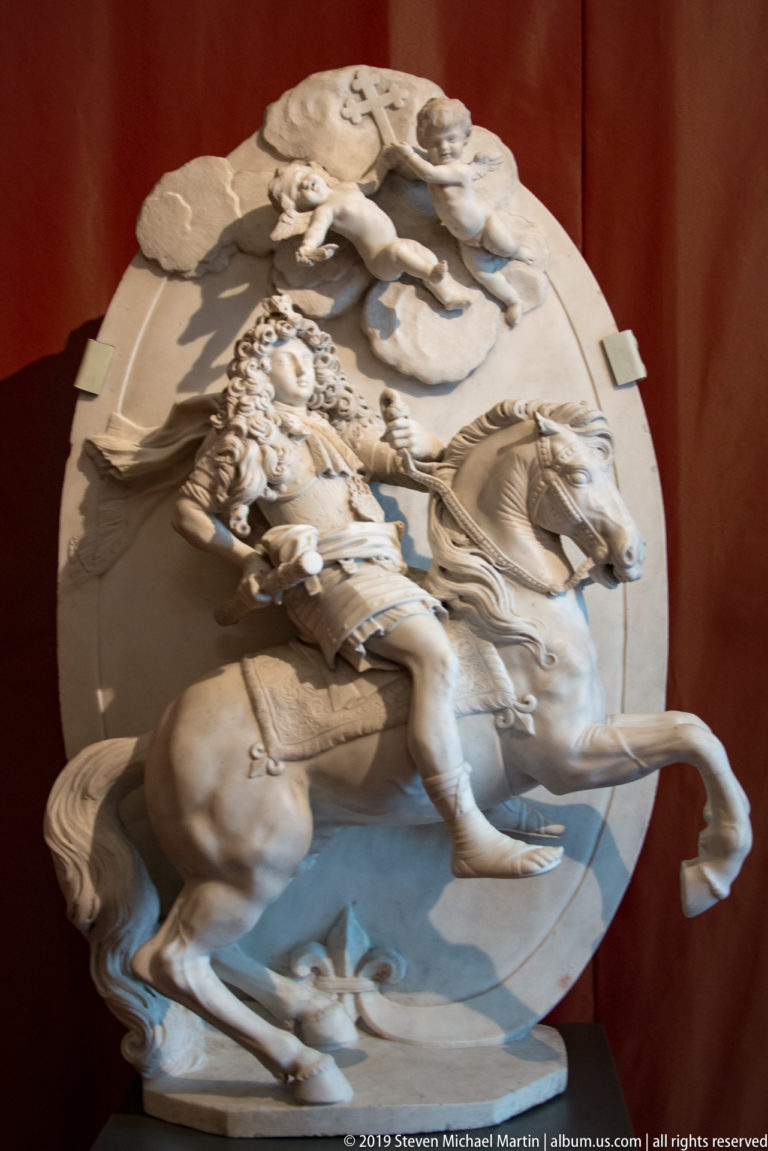 White relief sculpture of an armored rider on a rearing horse with additional figures above.