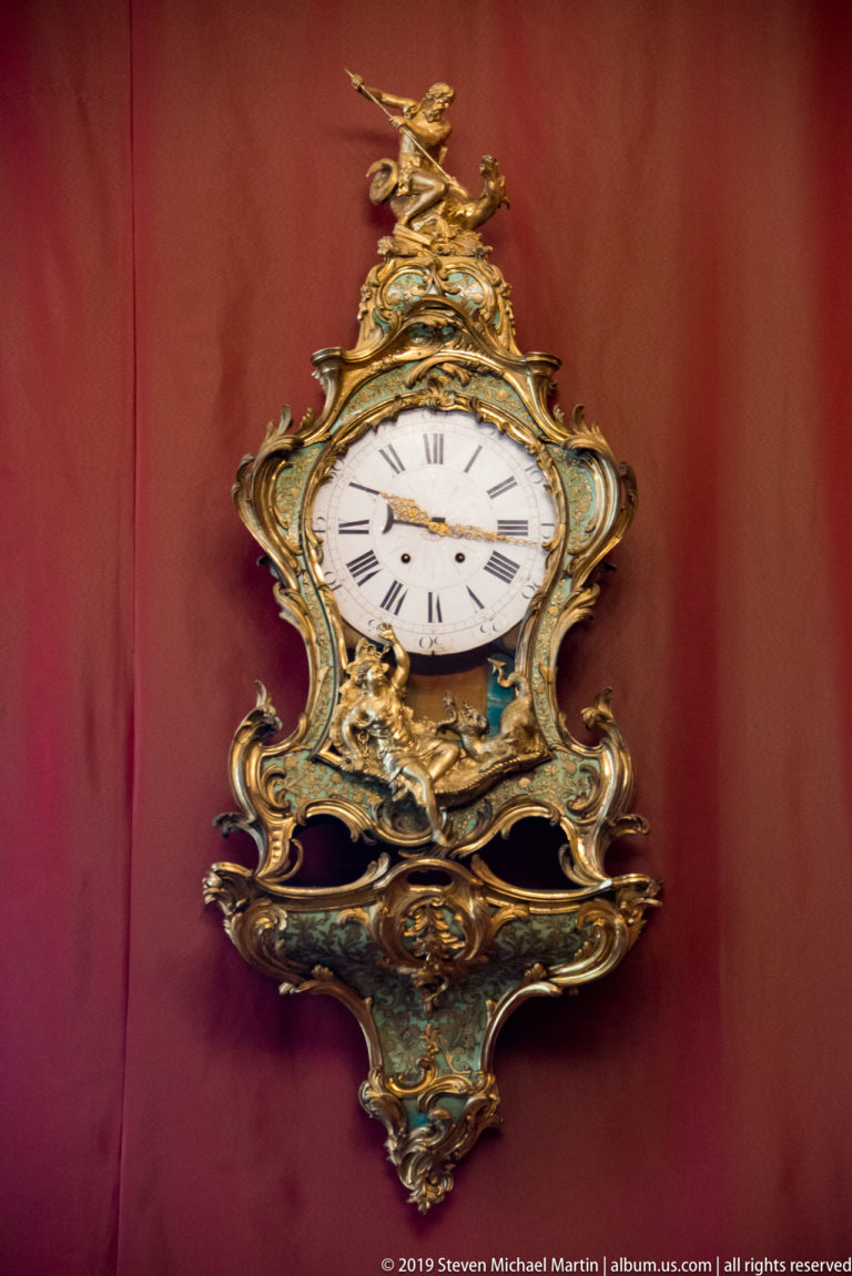 Ornate gilded clock with white Roman-numeral dial, blue-green painted panels, and sculptural crest.