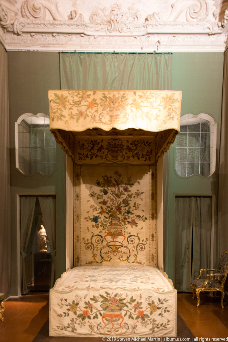 Canopy bed covered in pale floral embroidery, set against green wall hangings in a palace room.