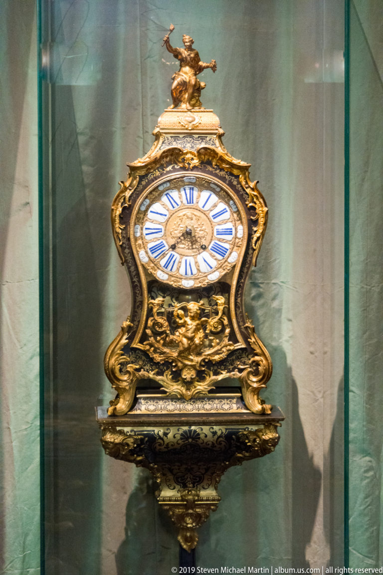 Ornate gilded wall clock with white dial, blue Roman numerals, scrolling case, and small figure at the top.