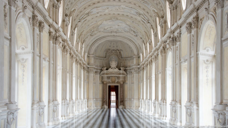 Axial view down the Galleria Grande toward an ornate end wall and doorway, with patterned floor and repeated windows.