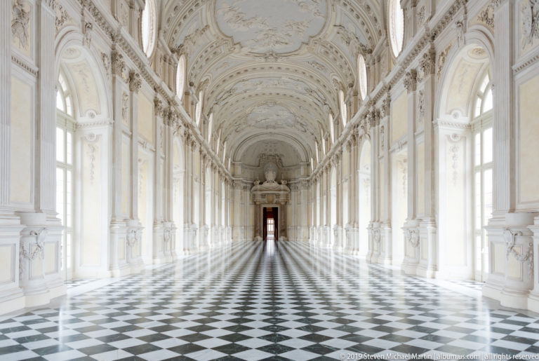 Centered symmetrical view through the Galleria Grande with arched windows, ornate white vault, and checker-pattern floor.