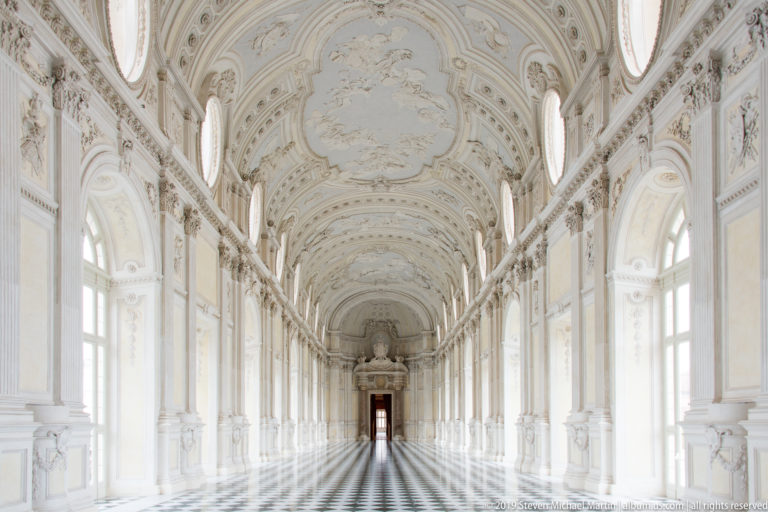 Long Galleria Grande seen from a slightly elevated angle, with ornate ceiling, arched windows, and patterned floor.