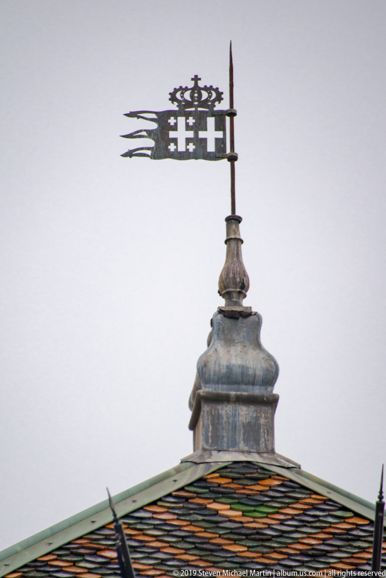 Metal heraldic weather vane above a roof patterned with green, yellow, and dark glazed tiles.