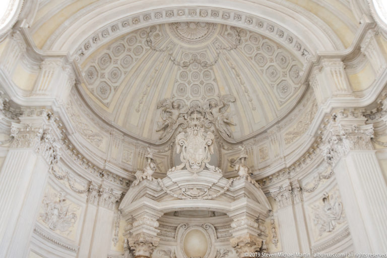 Large pale stucco cartouche beneath patterned vaulting and layered architectural moldings in the Galleria Grande.