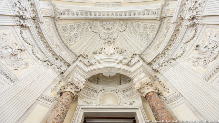 Upward view of ornate white vaulting, sculpted stucco, and marbleized columns above a doorway in the Galleria Grande.