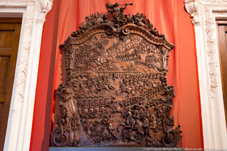 Large dark carved wooden relief packed with figures, horses, banners, and architectural scenes on a red wall.