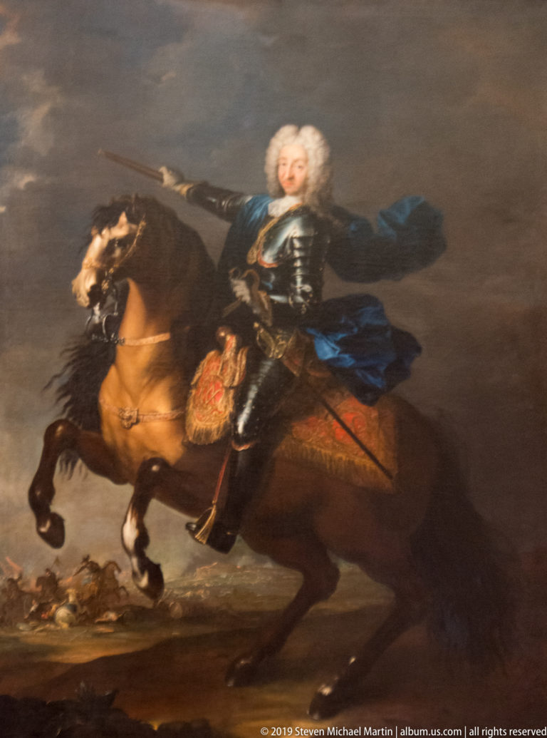 Painted aristocratic rider in armor and pale wig on a rearing brown horse against a dark landscape.