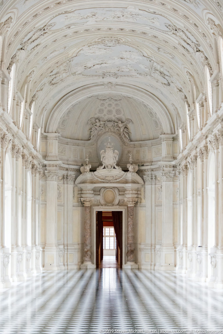 Symmetrical view down the empty Galleria Grande toward a monumental doorway beneath an ornate white vault.