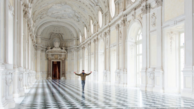 Visitor with arms outstretched in the Galleria Grande, surrounded by arched windows, white ornament, and a geometric floor.