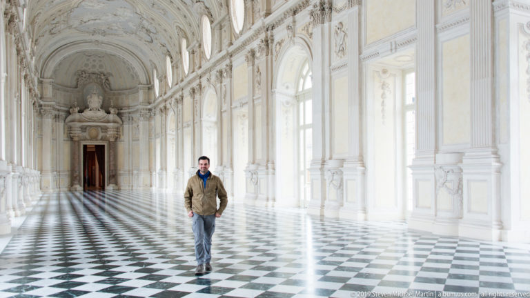 Man standing in the long white Galleria Grande with arched windows, ornate vault, and black-and-white geometric floor.