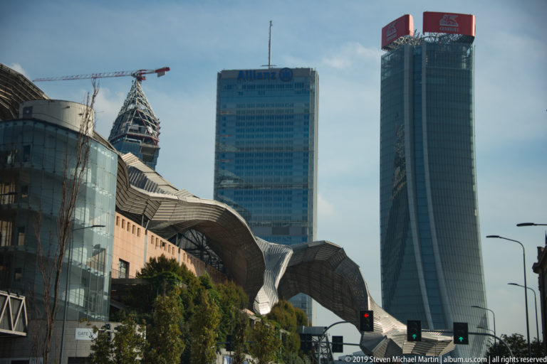 Modern Milan skyline with glass office towers and a large sculptural metal-roofed complex in the foreground.