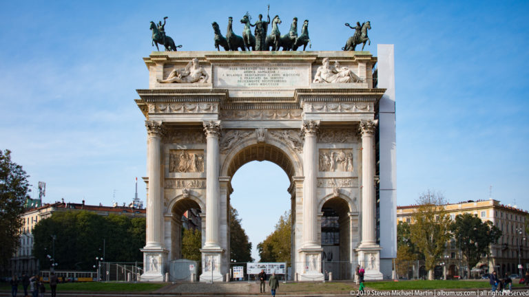 Frontal view of Milan’s Arco della Pace with three arches, Corinthian columns, reliefs, and bronze sculpture above.