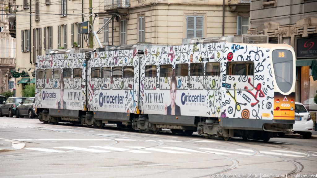 Articulated Milan tram on route 16 with a graphic advertising wrap, curving across tracks at a city intersection.
