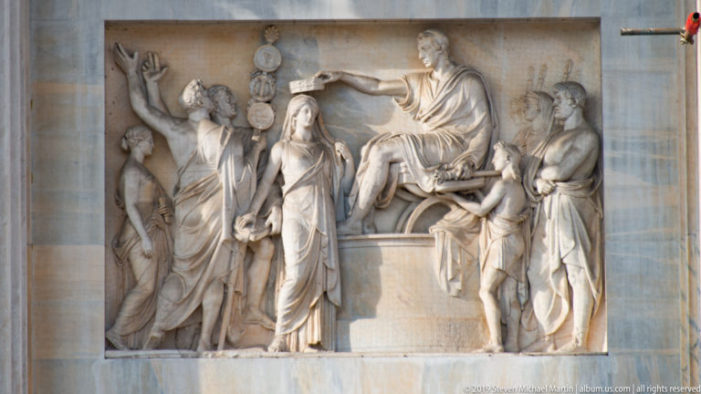 Marble relief on the Arco della Pace with a seated male figure, attendants, women, and circular emblems.