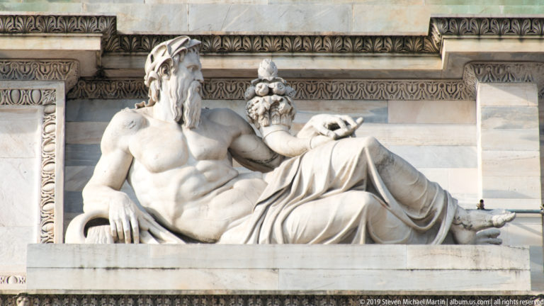 Reclining bearded marble figure beside a fruit-filled cornucopia on Milan’s Arco della Pace.