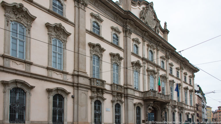 Oblique street view of Palazzo Litta’s ornate Baroque façade with tall windows, sculpted surrounds, balconies, and flags.