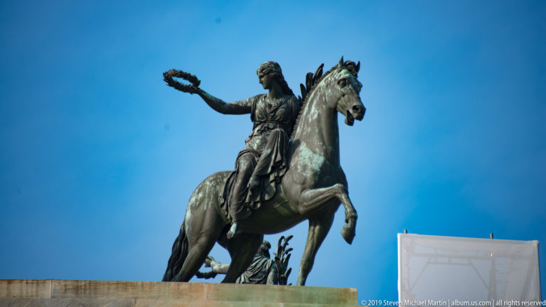Side view of a bronze female Victory on a rearing horse extending a wreath atop the Arco della Pace.