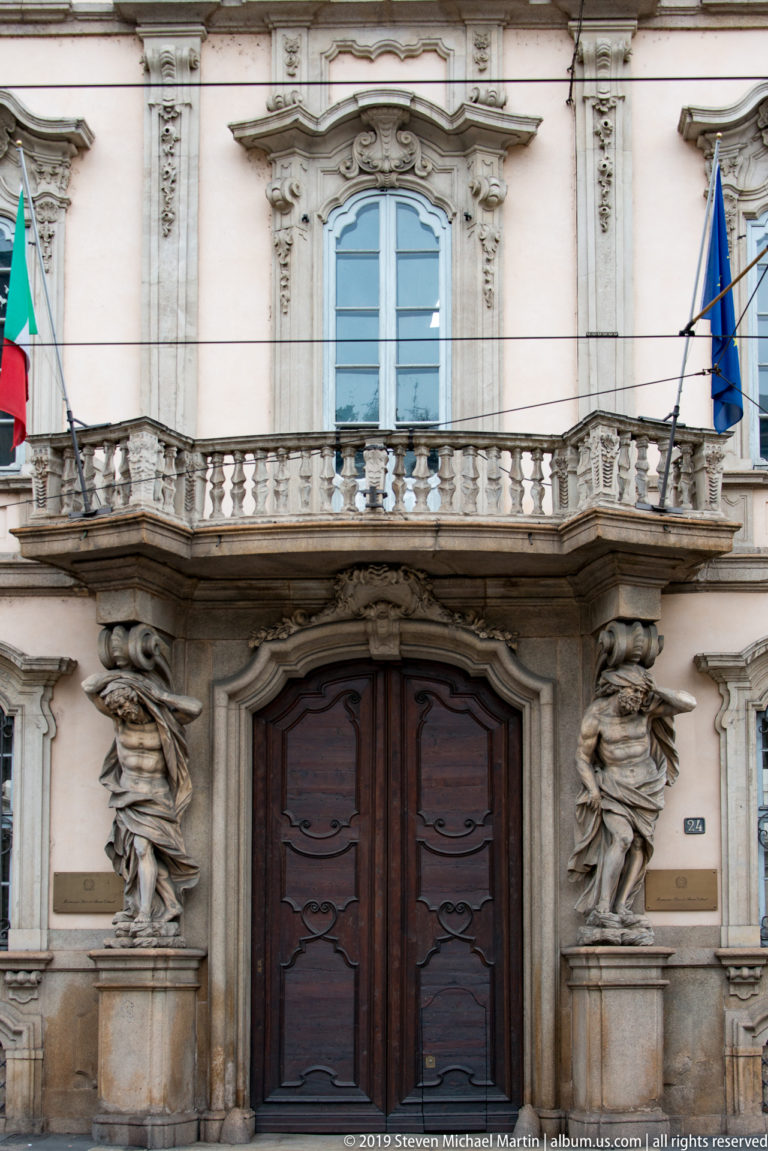 Ornate entrance of Palazzo Litta with a dark double door, sculpted atlantes, balcony, and Italian and EU flags above.
