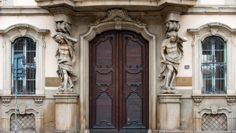 Baroque palace doorway in Milan flanked by sculpted atlantes, with wrought-iron window grilles on both sides.