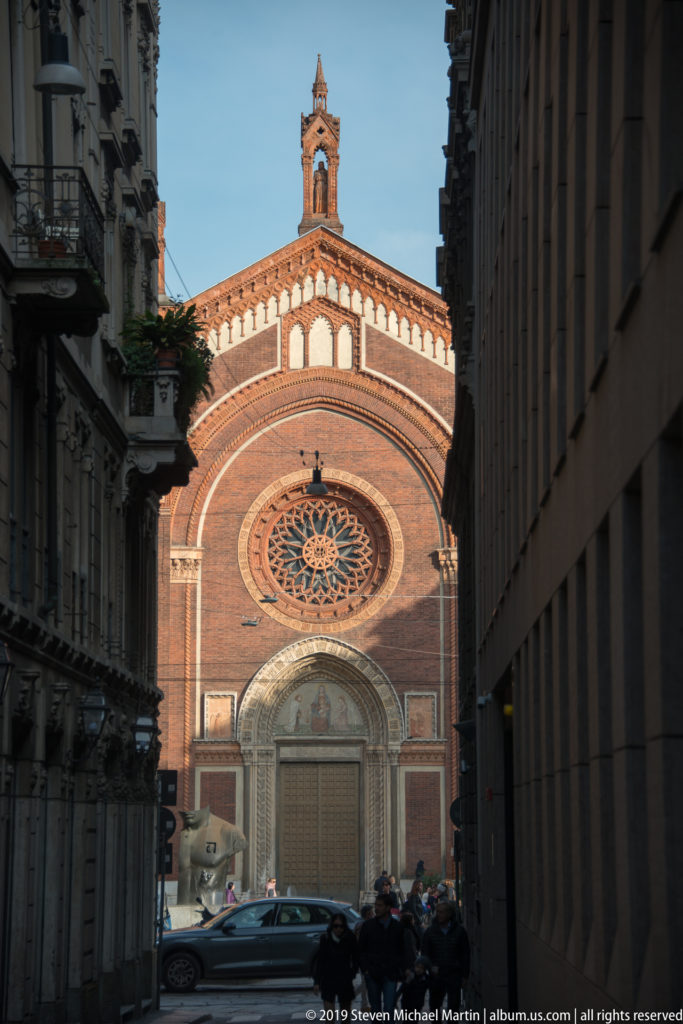 Brick church façade at the end of a narrow Milan street, with a large rose window, arched portal, and small bell gable.