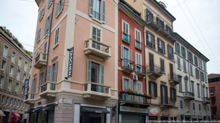 Narrow Milan street lined with pink, red, and gray residential façades, small balconies, shops, and overhead tram wires.