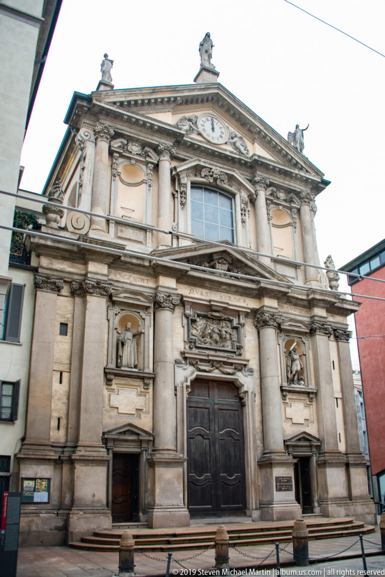 Baroque church façade in central Milan with a large wooden portal, paired columns, sculpture niches, and a triangular pediment.