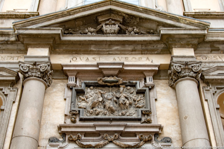 Baroque church façade detail with Corinthian columns, a carved relief panel, pediment, and Latin inscription.