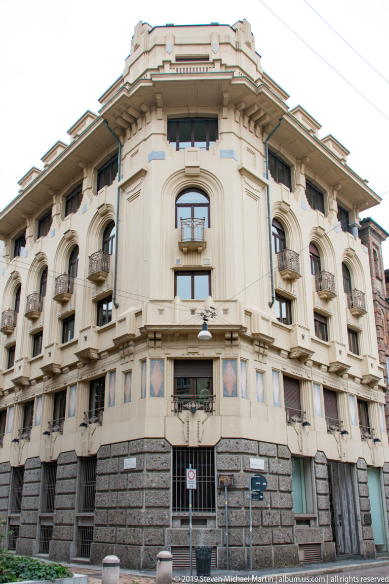 Curved corner apartment building with small balconies, arched upper windows, geometric panels, and a dark rusticated base.