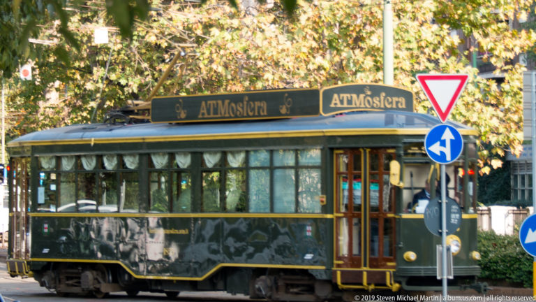 Dark green and gold ATMosfera dining tram on a Milan street, with large windows and its name on the roofline.