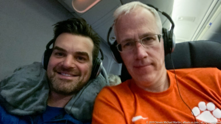 Two smiling male airline passengers seated together in an aircraft cabin and wearing over-ear headphones.