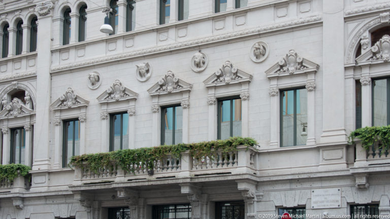 Pale stone façade with sculpted window pediments, round relief medallions, and a narrow balcony draped with greenery.