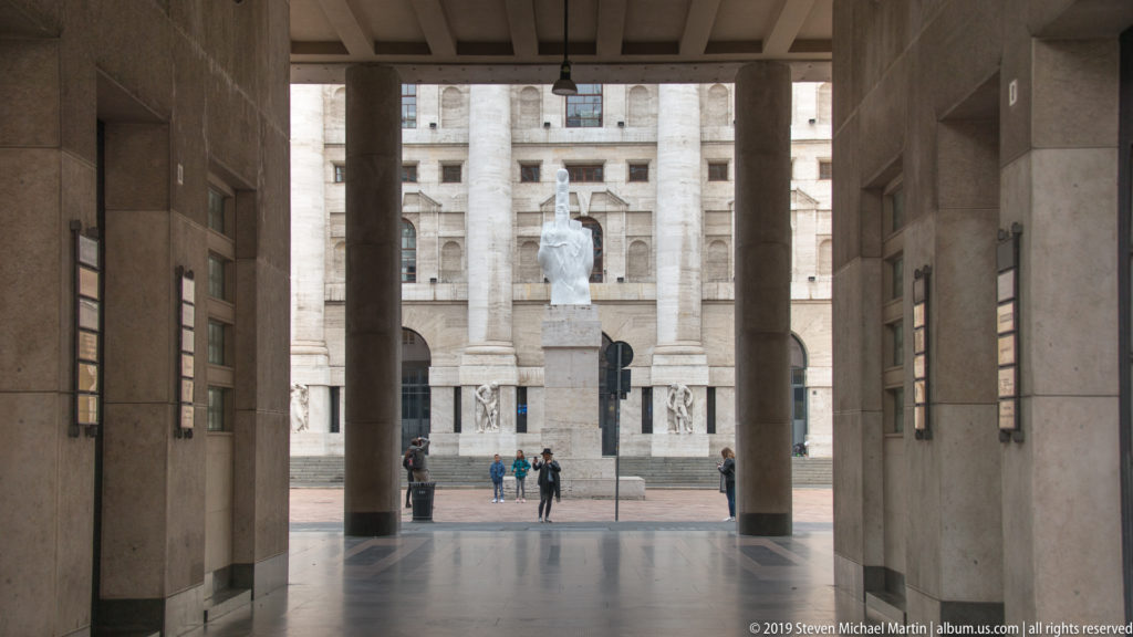 Maurizio Cattelan’s L.O.V.E. sculpture framed by dark columns, with Palazzo Mezzanotte across Piazza Affari.
