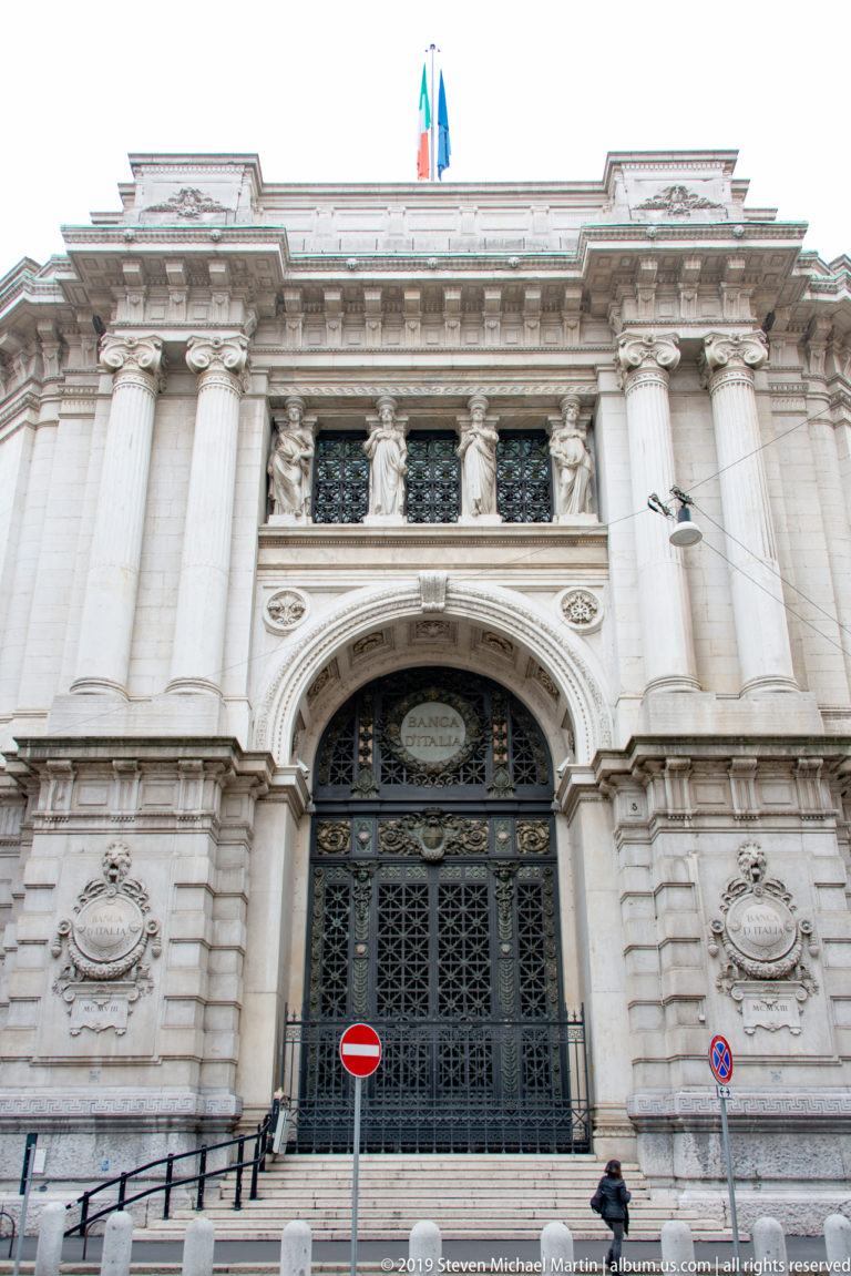 Grand stone institutional entrance with colossal columns, sculpted panels, ornate metal gates, and an Italian flag above.