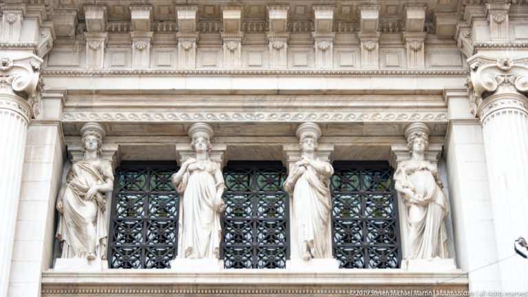 Four draped female stone figures standing between dark patterned window grilles beneath a classical entablature.