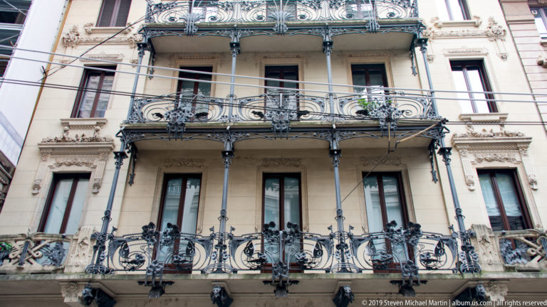 Pale Milan residential façade with stacked balconies enclosed by elaborate dark wrought-iron railings.