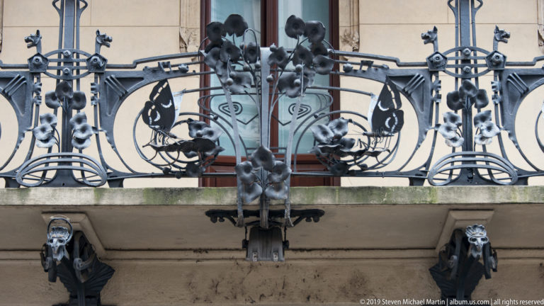 Ornate wrought-iron balcony with curling floral motifs, hanging forms, and grotesque masks beneath the ledge.