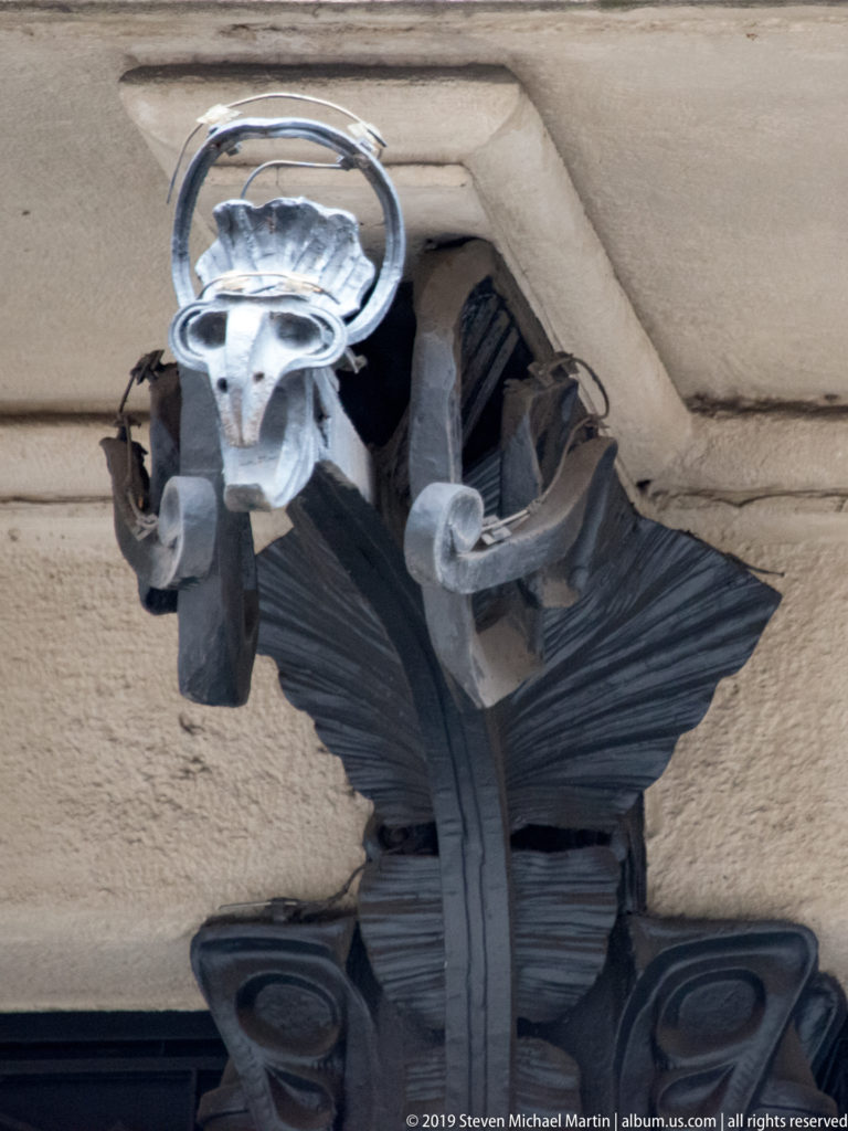 Close-up of a grotesque mask beneath layered black wrought-iron leaves and scrollwork on a Milan façade.