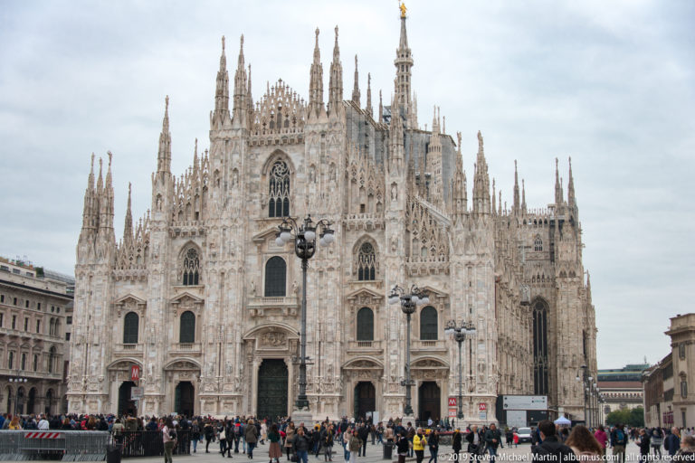 Frontal view of Milan Cathedral’s marble façade with five portals, dense Gothic sculpture, pinnacles, and crowds in the square.