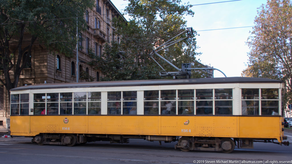 Long yellow vintage Milan tram crossing a city street beneath overhead electrical wires.