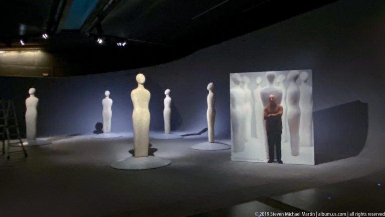 Dark gallery installation with multiple elongated pale human figures and a visitor standing inside a reflective enclosure.