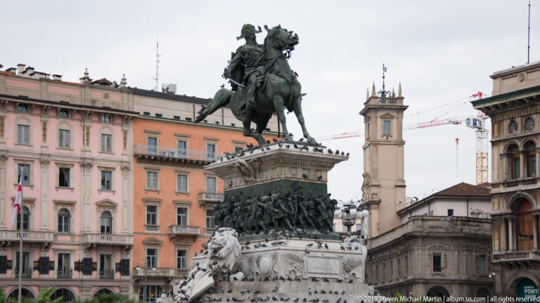 Oblique view of the Victor Emmanuel II equestrian monument with historic Piazza del Duomo façades behind.
