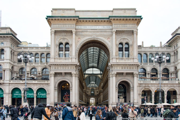 Symmetrical frontal view of the Galleria Vittorio Emanuele II entrance and glass-roofed arcade with crowds below.