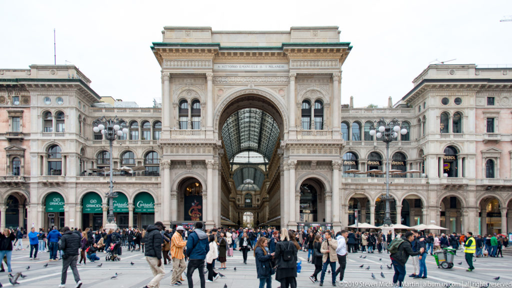 Wide view of the Galleria Vittorio Emanuele II entrance with crowds and surrounding arcaded buildings in Piazza del Duomo.