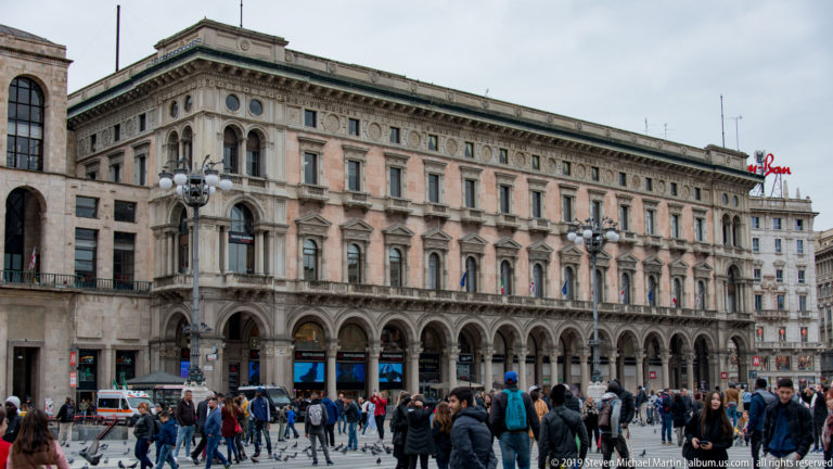 Long arcaded historic building on Piazza del Duomo with pedestrians crossing the square in front.